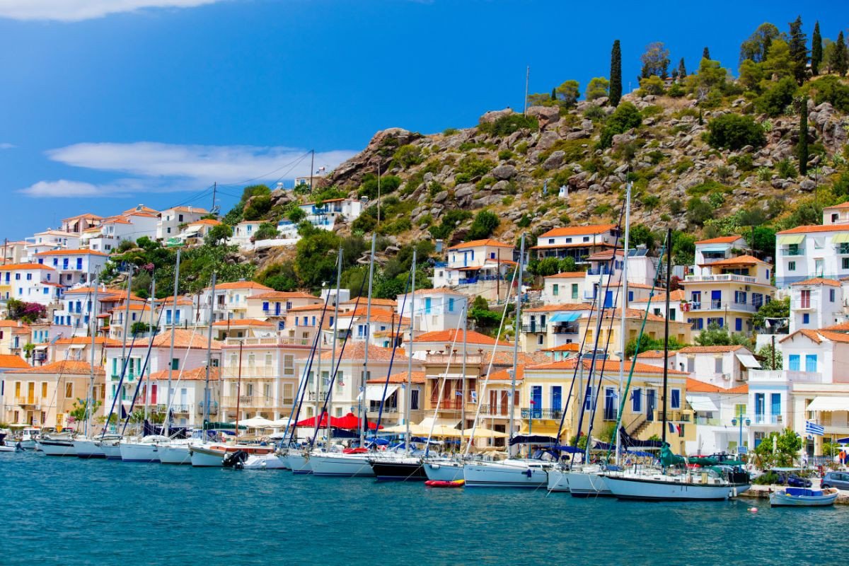 A picturesque view of a coastal town with colorful houses and sailboats docked along the waterfront, nestled against a rocky hillside under a clear blue sky.